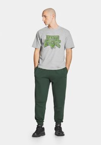 Szara bawełniana koszulka z zielonym nadrukiem "PROSTO ATHLETIC", w zestawie z ciemnozielonymi spodniami dresowymi i czarnymi butami sportowymi.