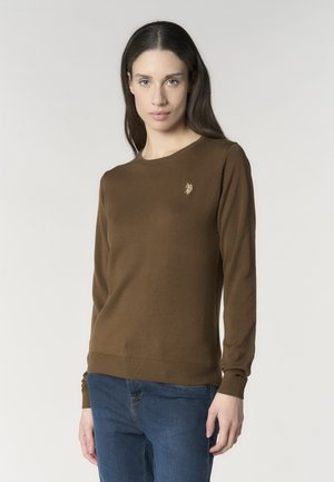 Donna con capelli lunghi e scuri che indossa un maglione marrone a maniche lunghe con un piccolo logo sul petto e jeans blu, rivolta in avanti su uno sfondo semplice.