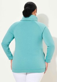 Pullover in pile color turchese con colletto alto, maniche lunghe a raglan e polsini a coste. Texture liscia con una vestibilità comoda, indossato sopra pantaloni bianchi.