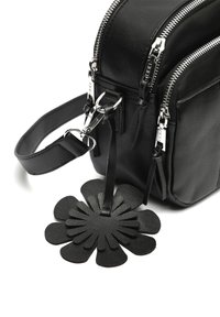 Schwarze Ledertasche mit zwei silbernen Reißverschlüssen an der Ecke, abnehmbarem Schulterriemen und schwarzem, blütenförmigem Schlüsselanhänger auf weißem Hintergrund.