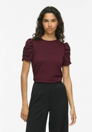 Femeie cu păr creț și închis la culoare, purtând un top texturat burgundy cu mâneci scurte bufante și pantaloni negri cu talie înaltă, stă într-un fundal alb.