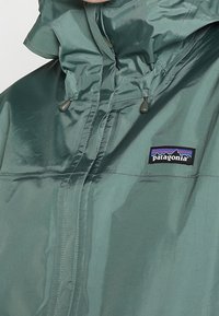 Grøn, vandtæt jakke lavet af skinnende materiale, med høj krave, justerbar hætte og et Patagonia-logo på brystet.