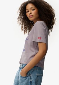 Camiseta de algodón color lila claro con mangas cortas, con gráficos de texto en azul y un pequeño parche de logo rojo en la manga izquierda. Corte holgado.