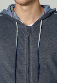 Sudadera con cremallera en tejido azul oscuro con acabado texturizado. Presenta un forro interior a rayas en azul claro y cordones grises.