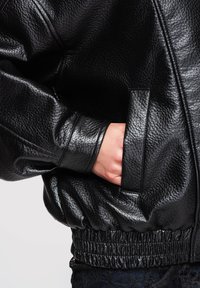 Mano metida en el bolsillo de una chaqueta de cuero negra texturizada con cintura y puño elásticos, sobre un fondo claro y liso.