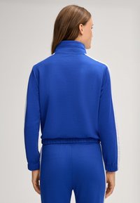 Felpa blu con colletto alto, texture morbida, polsini elasticizzati e strisce laterali bianche. Design aderente, abbinata a pantaloni blu coordinati.