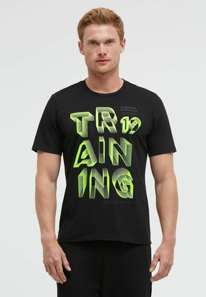 Man with light hair wearing black T-shirt with large green 3D text "TRAINING 19" standing against plain background.