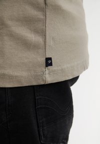 Zelené bavlněné tričko s lemem a malou modrou nášivkou. Nosit s černými džíny, které mají texturované prošití podél kapes.