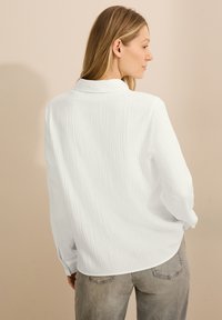 Hvid tekstureret skjorte med krave, lange ærmer og løs pasform, parret med lysegrå jeans. Enkel design, minimale detaljer.
