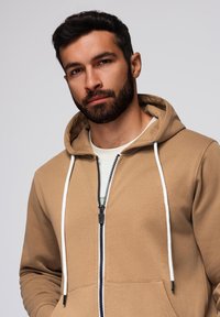 Felpa con zip color sabbia, dotata di cordini bianchi e una cerniera. Presenta una tasca a marsupio e un tessuto morbido e strutturato; indossata sopra una maglietta bianca.