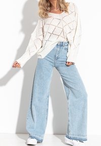 Weißer, offener Strickpullover mit Rautenmuster, kombiniert mit hellblauen Hochtaillieren und weitem Schnitt, sowie weißen Sneakers.