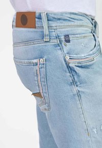 Jeans de mezclilla azul claro con un corte de altura media, parche de cuero marrón en la cintura y detalles de costura coloridos en los bolsillos. Elementos de diseño desgastados.