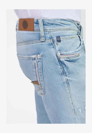 Jeans de mezclilla azul claro con un corte de altura media, parche de cuero marrón en la cintura y detalles de costura coloridos en los bolsillos. Elementos de diseño desgastados.