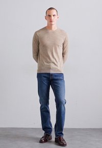 Pull en tricot beige avec un col rond, associé à un jean bleu et des chaussures en cuir marron foncé. L'ensemble présente un design simple et classique.