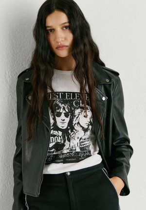 Giovane donna con lunghi capelli scuri indossa una maglietta bianca con stampa grafica e una giacca di pelle nera, in piedi davanti a una parete bianca e liscia.