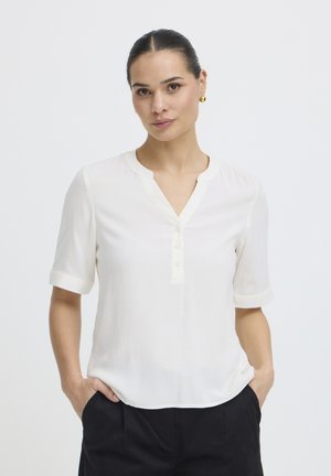 Kobieta z ciemnymi włosami związanymi do tyłu, ubrana w biały krótki rękaw z zapięciem na guziki i czarne spodnie, stojąca z rękami w kieszeniach na białym tle.