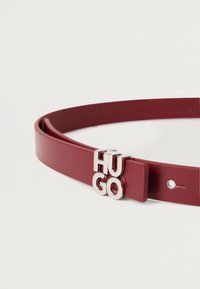 Ceinture en cuir rouge avec une boucle en métal argenté portant les lettres "HUGO." Texture lisse et design minimaliste, avec un seul trou d'ajustement.