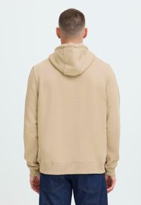 Uomo con capelli corti che indossa un hoodie beige e jeans blu scuro, girato di spalle su uno sfondo chiaro e semplice.