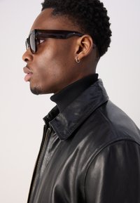 Μαύρο δερμάτινο μπουφάν με γιακά και λεπτομέρεια φερμουάρ, συνδυασμένο με στρογγυλά γυαλιά ηλίου και μαύρο ζιβάγκο. Απαλή υφή και κομψός σχεδιασμός.