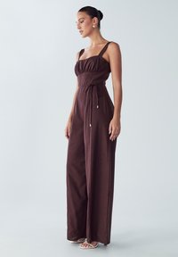 Mono burdeos con piernas anchas, corsé ajustado y cinta ajustable en la cintura. Presenta tirantes gruesos y una tela ligera y suave.
