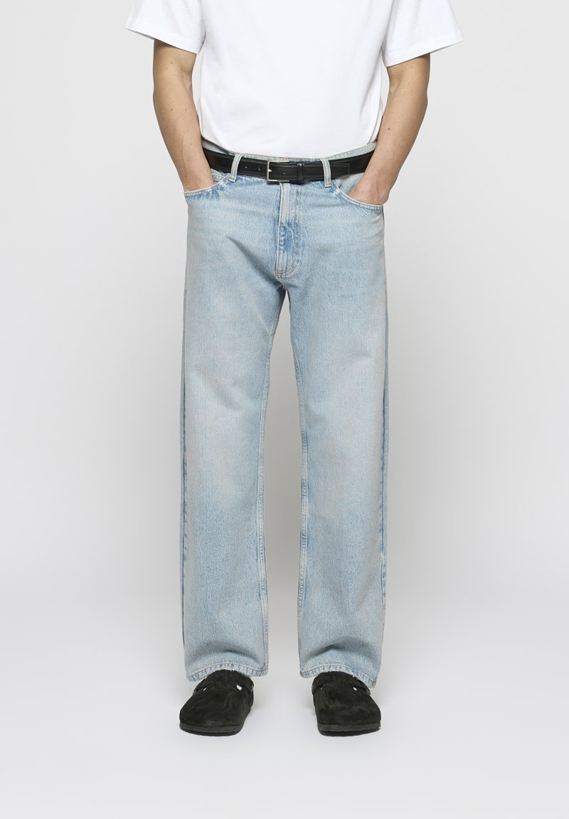 Jeans de ganga azul claro com perna reta, design de cinco bolsos e um cinto preto. Usados com uma t-shirt branca lisa e sapatos pretos.