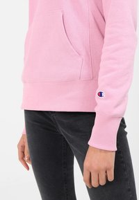 Lichtroze hoodie met een voorkraag, ribbelboorden en -zoom; heeft een klein geborduurd logo op de mouw. Het materiaal lijkt zacht en textuur te hebben.