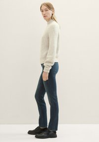 Suéter beige de canalé con un cuello alto, combinado con jeans ajustados azul oscuro y zapatos negros con cordones, mostrado de lado contra un fondo neutro.