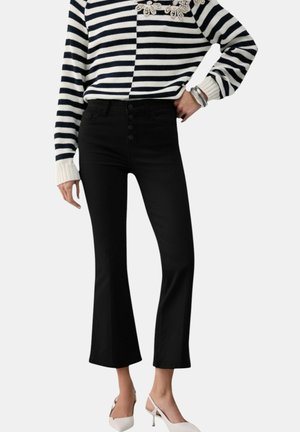 Donna che indossa pantaloni neri a zampa a vita alta, maglione a righe bianche e nere, décolleté bianche con cinturino posteriore e braccialetti d'argento al polso.