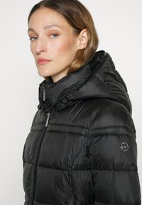 Schwarze Daunenjacke mit Kapuze, ausgestattet mit einem Reißverschluss vorne, gestepptem Design und einem Logopatch am linken Arm. Glattes, glänzendes Material.