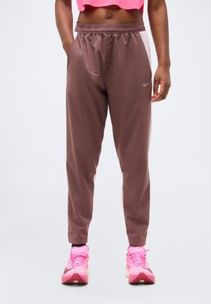 Γυναίκα που φοράει καφέ παντελόνι της Nike με ελαστική μέση και τσέπες, συνδυασμένο με φωτεινά ροζ αθλητικά παπούτσια της Nike και ένα κοντομάνικο ροζ τοπ.