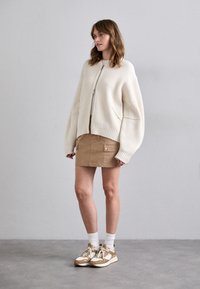 Kremfarget ribbstrikket cardigan med glidelås foran, sammen med et brunt miniskjørt med lommer og gullakenter, samt beige treningssko.