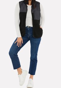 Chaleco de forro polar negro con hombros acolchados grises y bolsillo en el pecho, llevado sobre una camisa blanca de manga larga, con jeans rectos azules y zapatillas blancas.