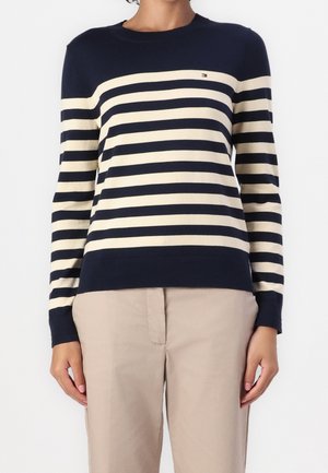 Kvinde iført en marineblå og cremefarvet langærmet sweater med vandrette striber og beige bukser mod en ensfarvet baggrund.