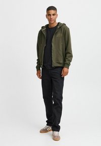 Felpa verde oliva con zip e coulisse, due tasche frontali e dettaglio del logo sul petto, abbinata a una maglietta nera, pantaloni scuri e sneakers.