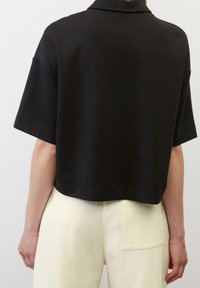 Černá krátká halenka s krátkým rukávem a límečkem, oblečená s vysokými off-white kalhotami s kapsou na zadní straně, viděná zezadu.