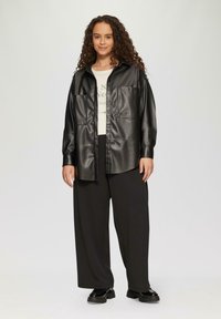Zwarte faux leren overhemdjack met grote zakken en een knoopsluiting aan de voorkant, gecombineerd met wijde zwarte broeken en een lichtgekleurde blouse eronder.