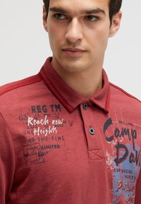Rotes Poloshirt aus Baumwollmischung mit strukturierter Oberfläche, zwei Knöpfen und bedruckten Grafiken mit Text auf der Vorderseite.