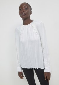 Blusa bianca plissettata con maniche lunghe, caratterizzata da un colletto tondo e maniche con polsini, realizzata in un tessuto lucido e morbido.