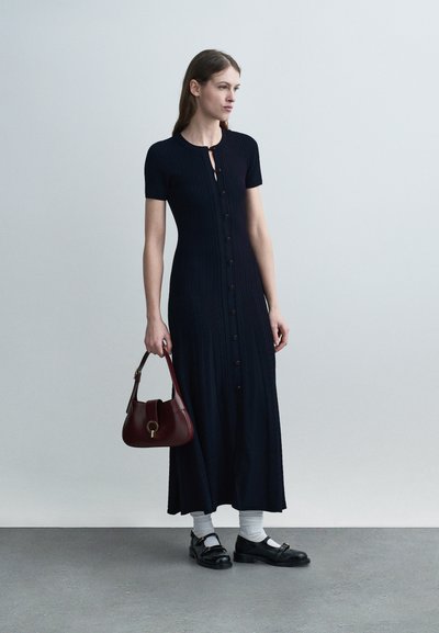 Γυναίκα με μαύρο midi φόρεμα με κουμπιά, κρατώντας μια μπορντό τσάντα, φορώντας λευκές κάλτσες και μαύρα παπούτσια Mary Jane, στέκεται σε γκρι πάτωμα.