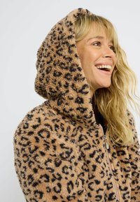 Sweat à capuche en peluche imprimé léopard aux tons doux de beige et noir, doté d'une grande capuche et d'une fermeture éclair. Tissu texturé et confortable.