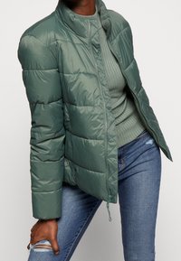 Giacca verde imbottita con colletto alto, chiusura con zip e finitura texturizzata. Indossata sopra un maglione a coste abbinato e jeans blu.
