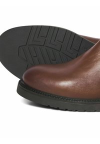 Bottes en cuir marron avec un bout arrondi, dotées d'une semelle en caoutchouc noir texturé et de détails cousus le long du bord.