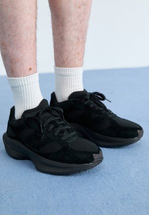 Person trägt schwarze Mesh- und Wildledersneaker mit dicken Sohlen und weißen gerippten Socken auf einem hellblauen Teppichboden.