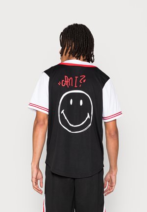 Personne avec des dreadlocks vue de dos, portant un t-shirt noir et blanc avec un liseré rouge, avec un texte rouge "can I?" et un grand smiley blanc dans le dos.