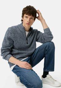 Jonge man op zijn knieën, gekleed in een grijze half-zip trui, blauwe spijkerbroek, zwarte sokken en witte sneakers, met één hand op zijn hoofd.