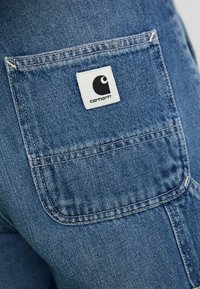 Džínová nohavica s vreckom a nápisom značky Carhartt šitým na zadnej strane, zobrazujúca detailné prešívanie a textúru látky.