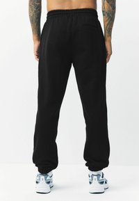 Pantalones de chándal negros con cintura elástica, puños ajustados y un bolsillo trasero. Hechos de una tela suave, con una textura lisa.