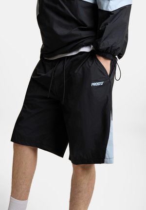 Osoba nosząca czarne szorty Prosto z jasnoniebieskimi bocznymi panelami i rękami w kieszeniach, w połączeniu z pasującą czarno-jasnoniebieską kurtką.