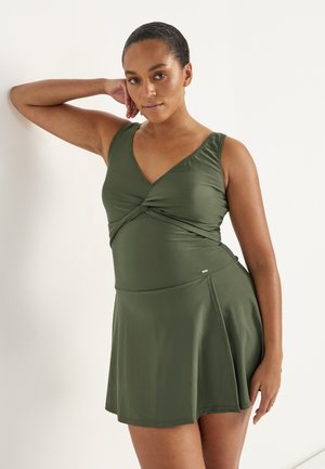 Vrouw staand met hand steunend op het hoofd, draagt een mouwloze olijfgroene jurk met V-hals en uitlopende rok.