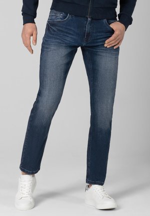Man draagt een slim-fit vervaagde blauwe spijkerbroek en witte sneakers, staat met één hand in de zak en de andere ontspannen langs het lichaam.